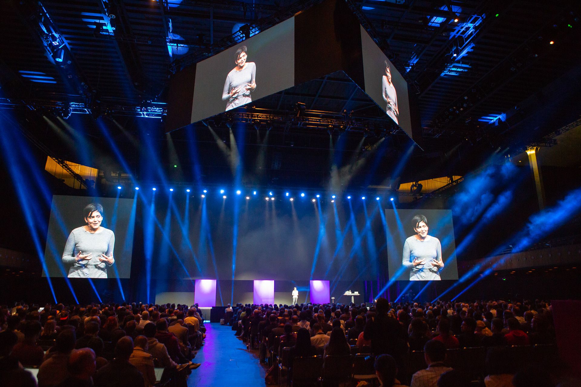 Large keynote hall with dramatic beam lighting and speaker displayed on multiple IMAG screens